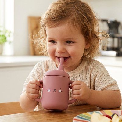Fillette souriante à table buvant dans son gobelet bebe rose